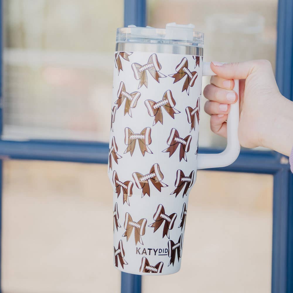 A white tumbler with a pattern of brown and black football bows held in a hand.